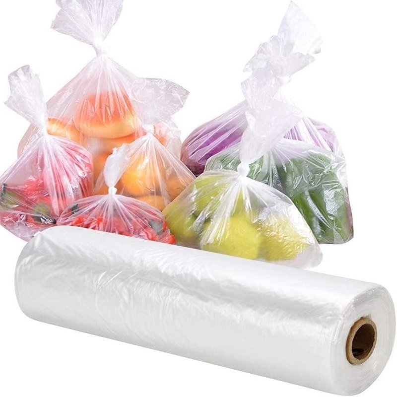 A roll of white biodegradable fruit and vegetable bags is shown in the foreground, and four transparent bags in the background contain colorful candies and fruits, demonstrating the product's form and usage scenarios.