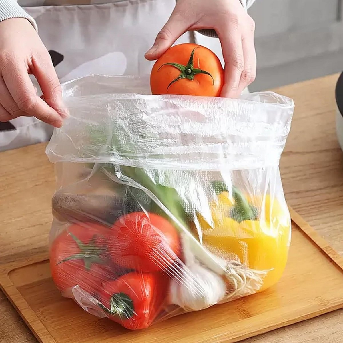 Hands are putting tomatoes into a transparent biodegradable fruit and vegetable bag, which contains a variety of fruits and vegetables, demonstrating the product's fresh-keeping usage scenario.